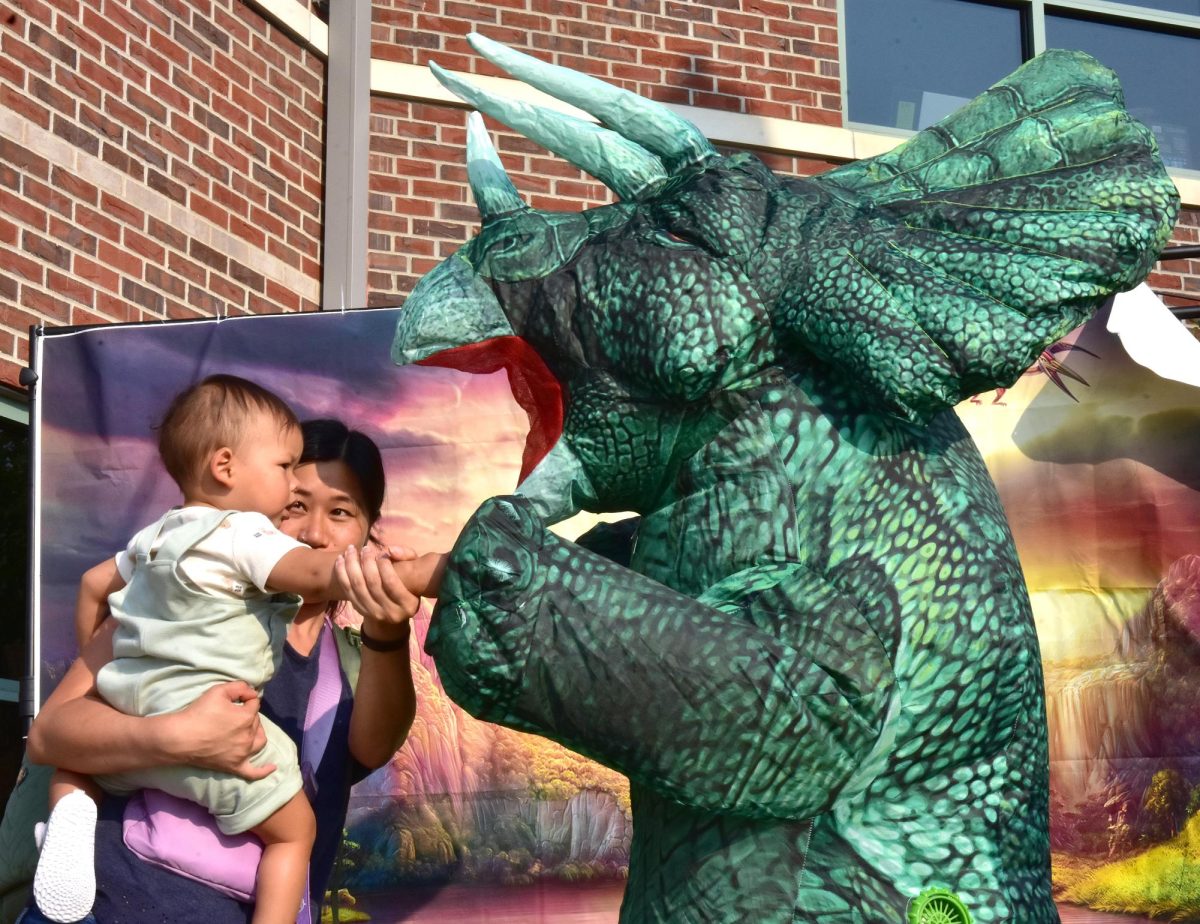 Xiang helps RJ fist-bump Trixie the Tyrannosaurus during the 4th annual Dinosaurs at Dusk event on the patio at the Solon Public Library. The event celebrated the life of Jack Neuzil, who hand-crafted well over 100 wooden dinosaurs combining his love of woodworking, dinosaurs, and improving the lives of children.